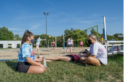 Two women sit and talk in front of an outdoor volleyball game while wearing masks