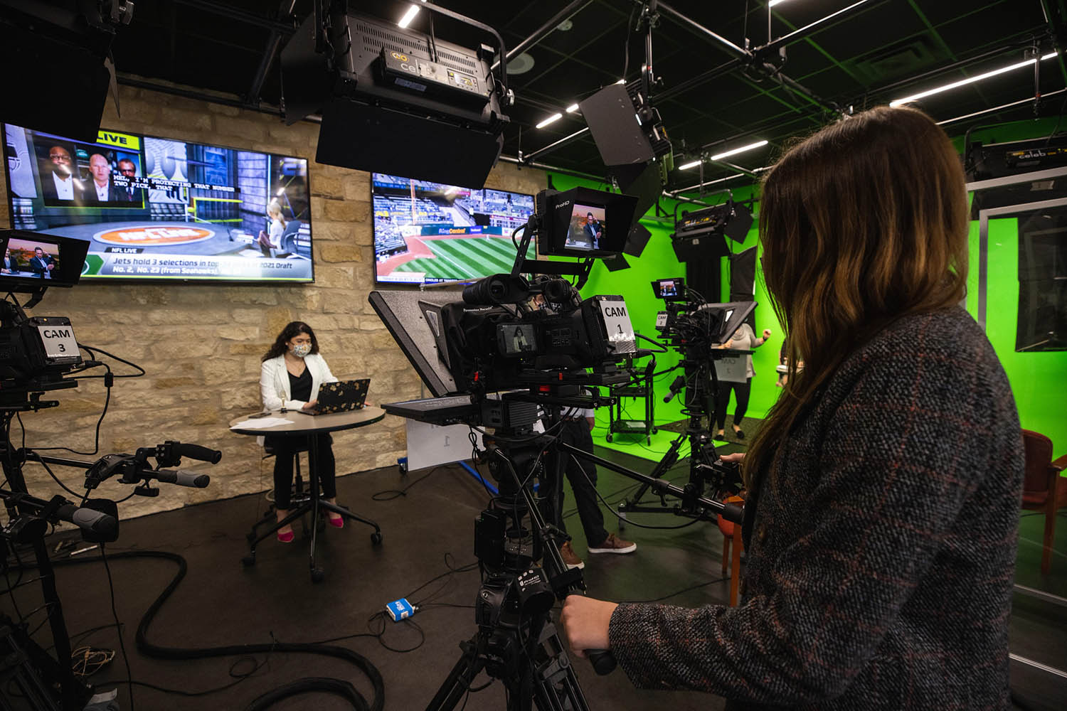 Student sitting at a table reading a laptop in a studio in front of video camera with TV screens in the background